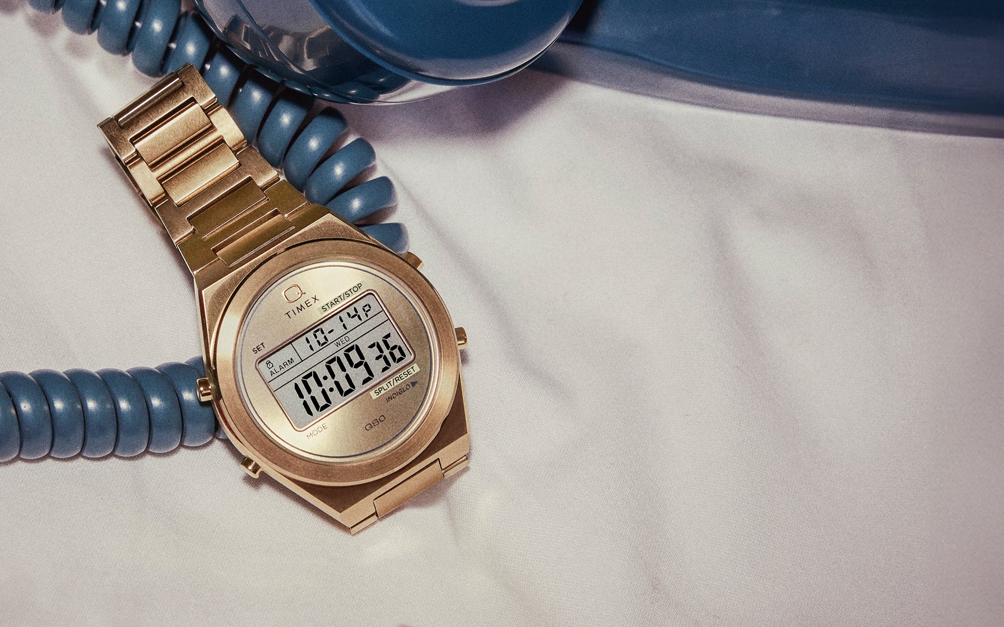 A gold-tone Q Timex® Q80 Continental digital watch resting on a white bedsheet, wrapped in the coiled cord of a vintage blue rotary telephone.