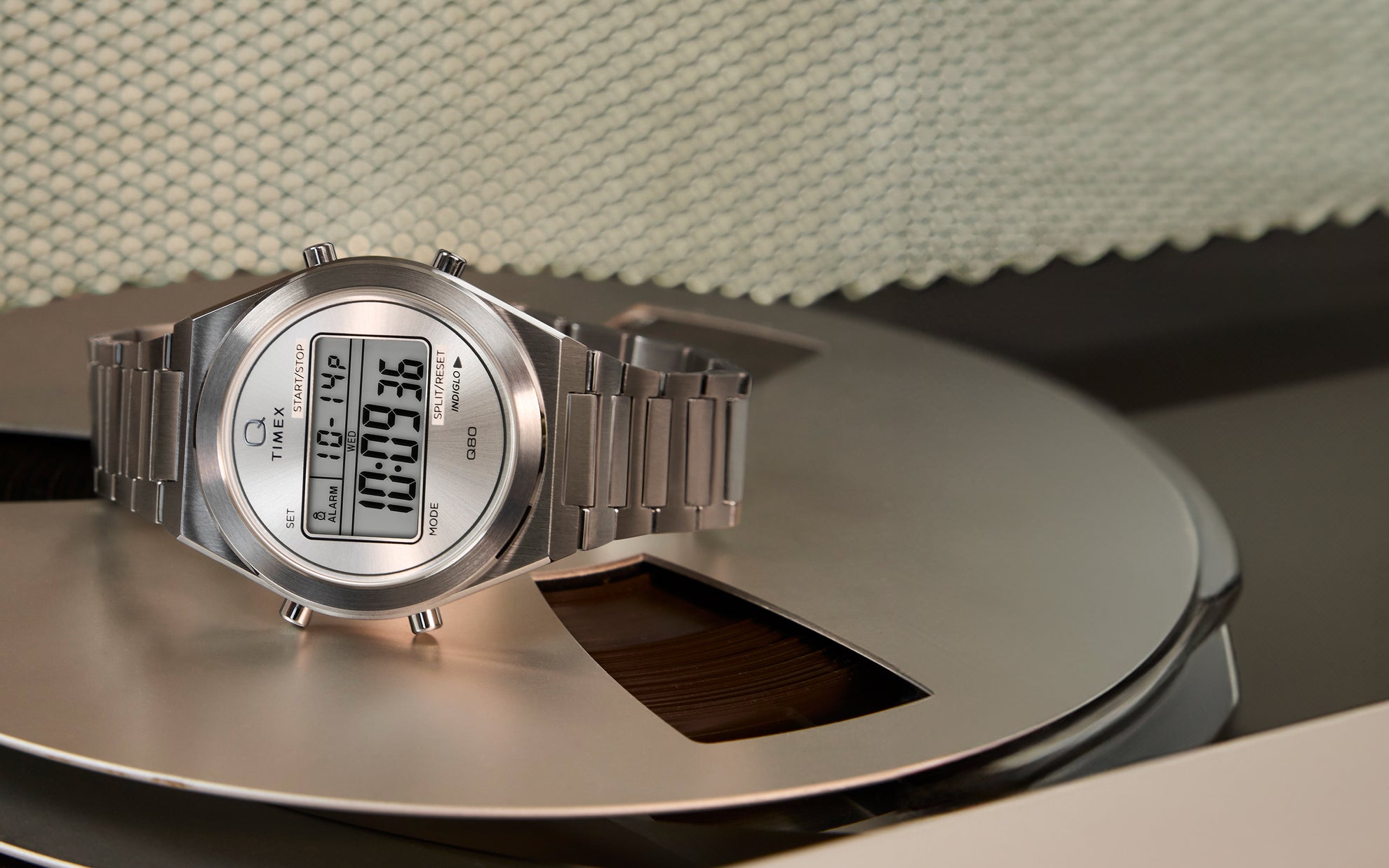 A gold-tone Q Timex® Q80 Continental digital watch with a round case and metal bracelet, displayed on a coiled blue telephone cord atop a white fabric surface. The digital display shows the time, day, and date.