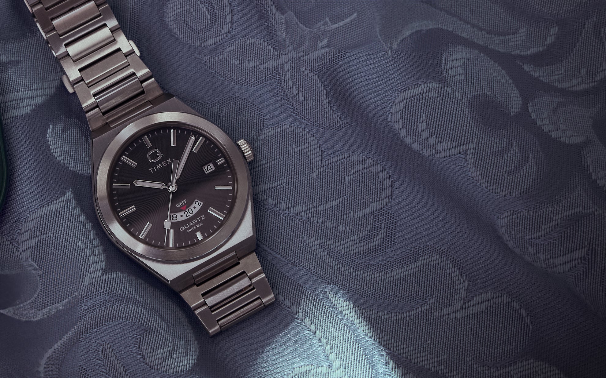 Q Timex® Continental GMT watch with a stainless steel case and bracelet, black dial, and red GMT hand, displayed on a textured navy blue fabric with floral jacquard pattern, showcasing its vintage-inspired design.