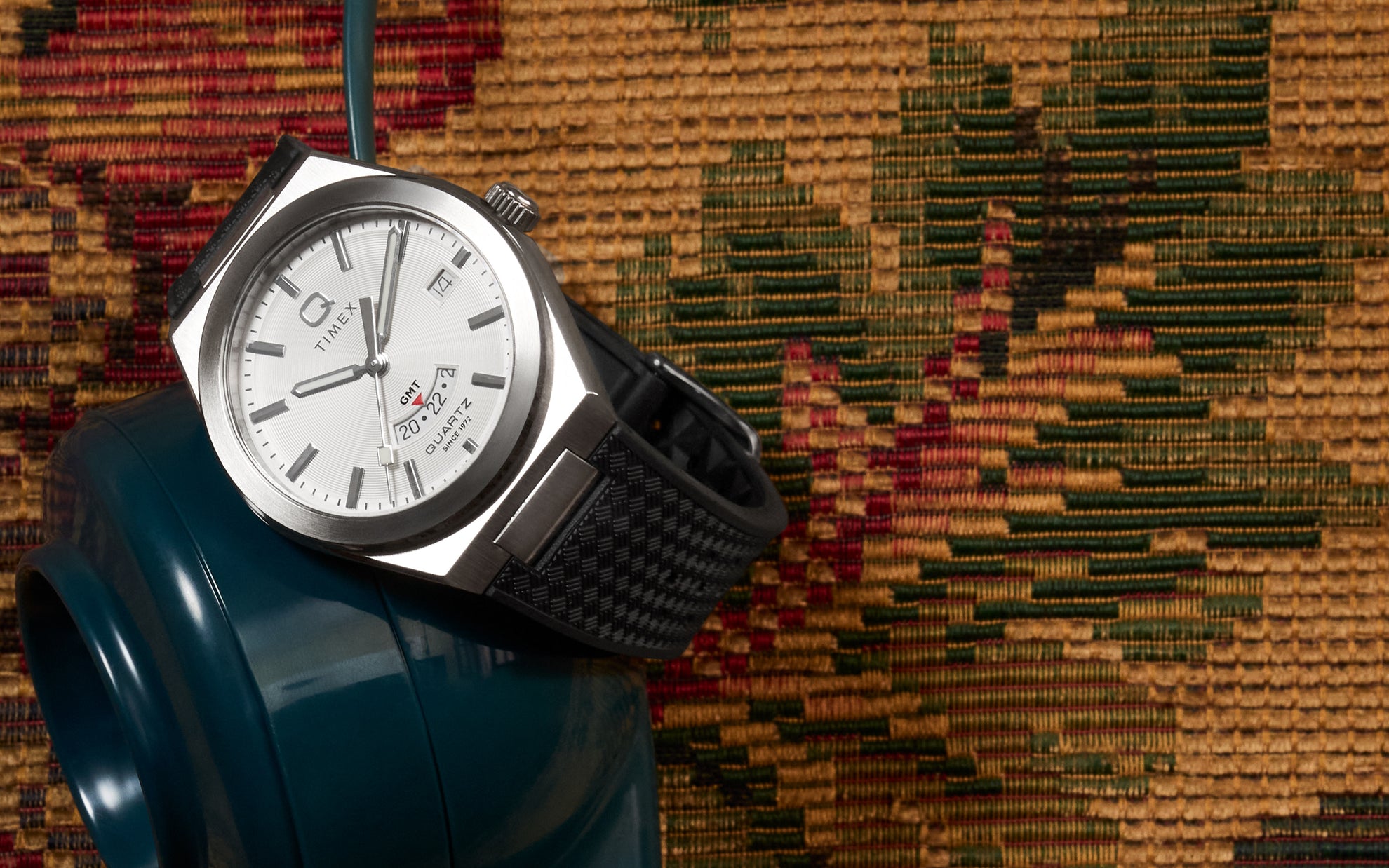 Q Timex® Continental GMT watch with a silver dial and black woven-texture strap, draped over a vintage teal rotary phone, with a warm tapestry textile in the background.