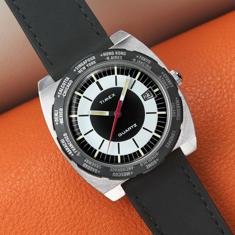 Timex Q Reissue world time watch with a two-tone black and white dial. Cities from around the world are printed on the rotating bezel. Set on a leather strap, styled against an orange textured surface.