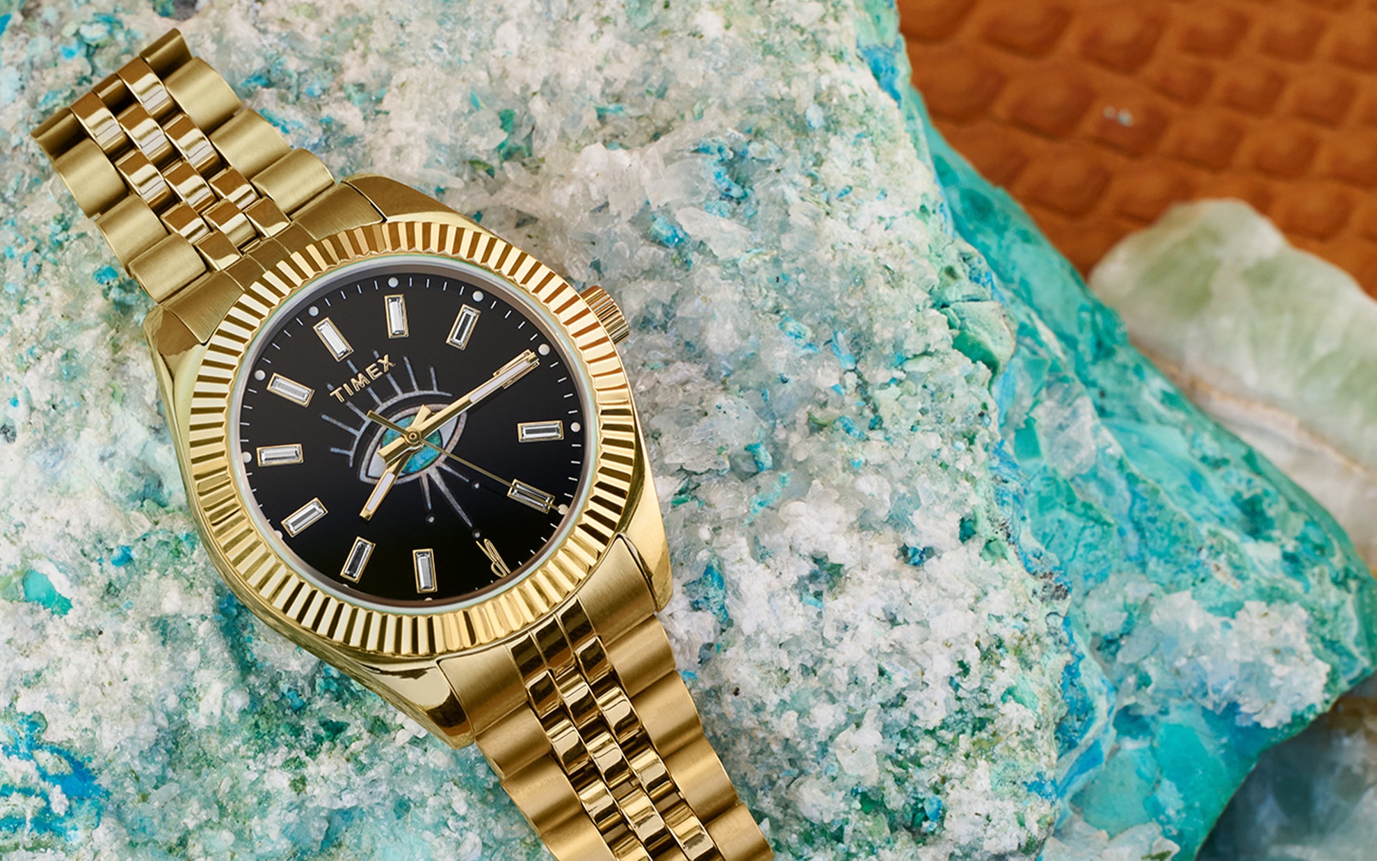 Timex x Jacquie Aiche gold-tone watch placed on textured blue and white mineral crystals. The timepiece features a polished gold bracelet and case, a fluted bezel, and a striking black dial with a central eye motif and baguette crystal hour markers.