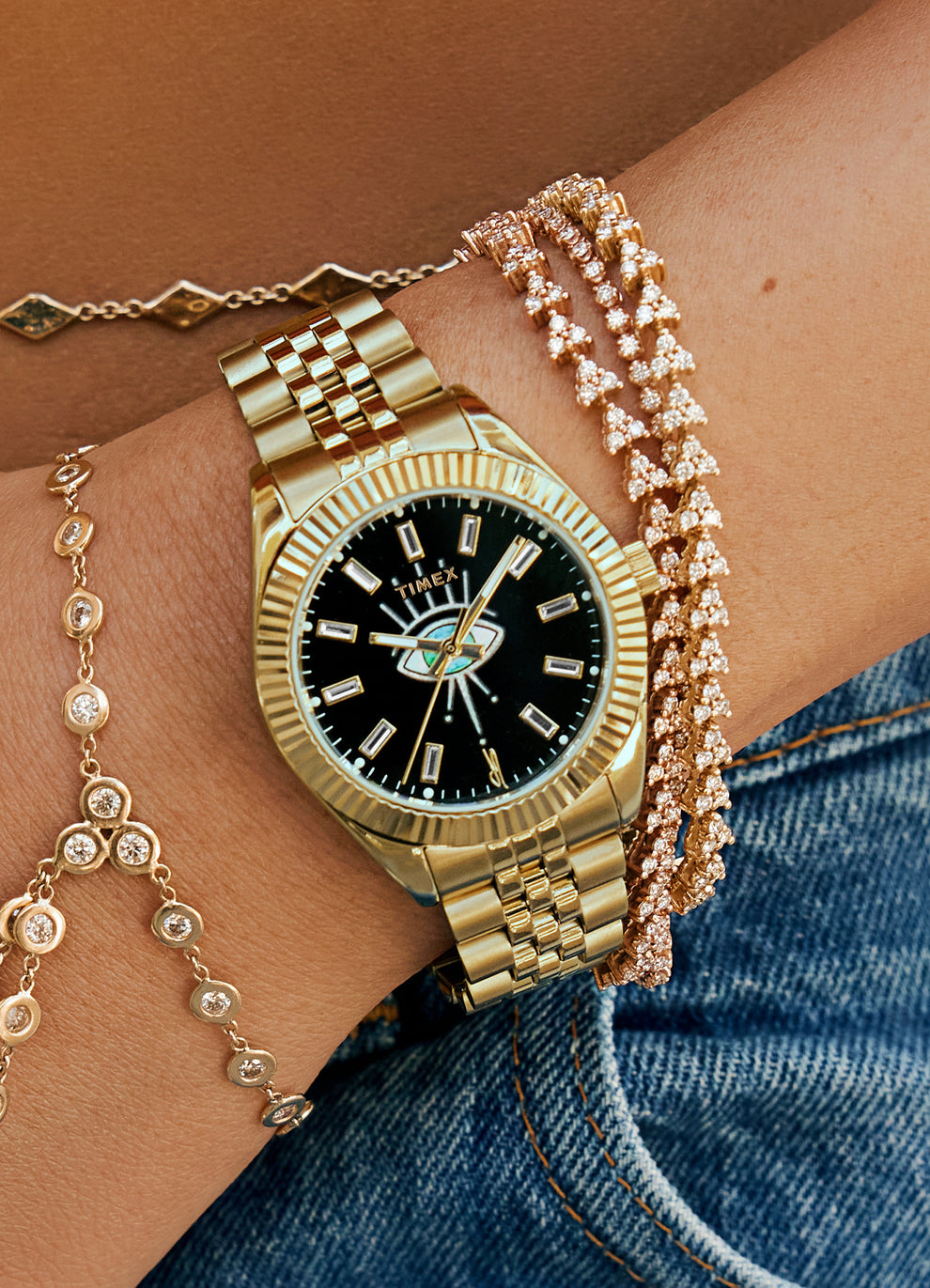 Timex x Jacquie Aiche gold watch styled on a wrist adorned with gold and crystal-studded bracelets. The watch has a black dial with a signature eye illustration, fluted gold bezel, and matching gold bracelet, worn against warm-toned skin and denim fabric.