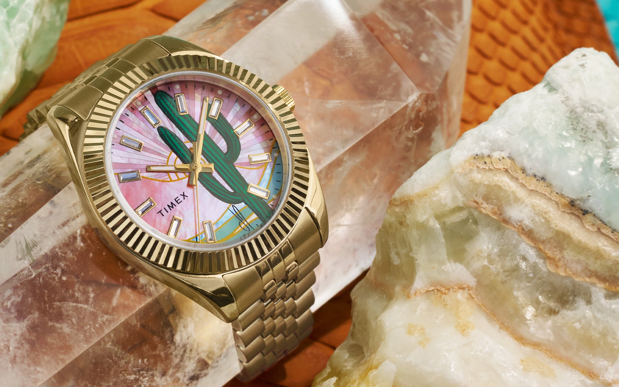 Close-up of a Timex x Jacquie Aiche gold-tone wristwatch placed on a rose quartz crystal, surrounded by other mineral stones. The watch face features a green cactus illustration against a pink mother-of-pearl dial with gold-tone hour markers and hands, encased in a fluted bezel and gold metal bracelet.