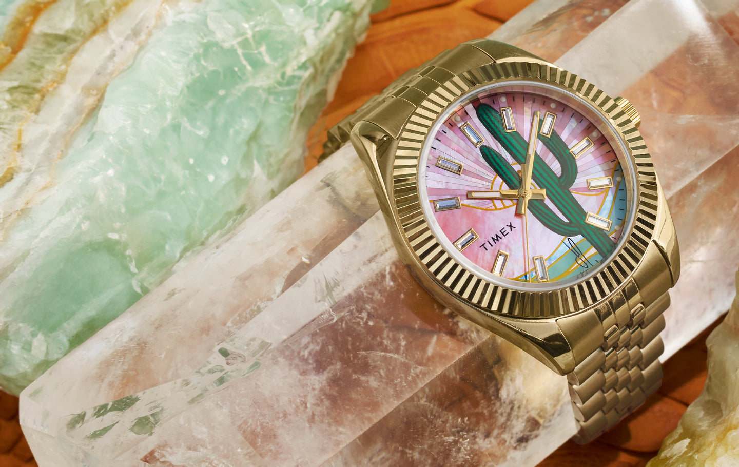 Gold Timex watch from the Jacquie Aiche Cactus collection, featuring a vibrant dial with a green cactus design on a pink and sunburst background, set on a clear crystal surface with green and amber mineral stones in the background.