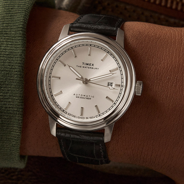Timex Waterbury Automatic watch with a silver sunray dial, stainless steel case, and black leather strap, shown on a wrist. The dial features silver-tone hour markers, date window at 3 o’clock, and text reading 'Established in 1854' at the bottom.