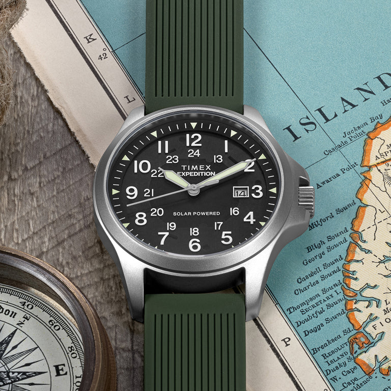 Timex Expedition Solar Powered watch with a black dial, luminous hands, silver-tone case, and green textured strap. The dial features both 12-hour and 24-hour markers along with a date window at 3 o’clock. The watch is placed on a vintage map with a compass, highlighting its rugged outdoor adventure style.