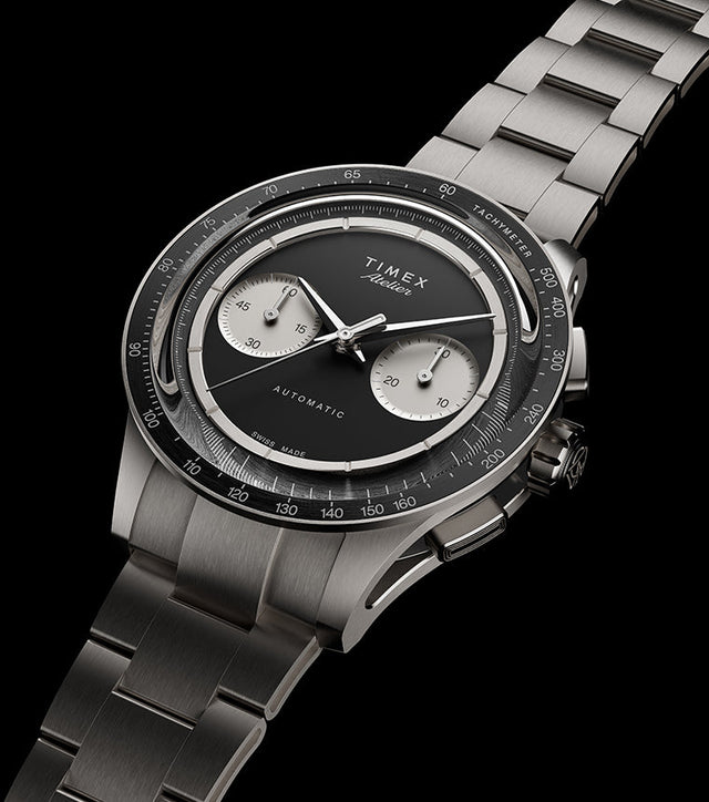 Timex Atelier Chronograph Automatic M1a Ti titanium watch with black dial, twin silver sub-dials, tachymeter bezel, and integrated titanium bracelet shown at an angled perspective.
