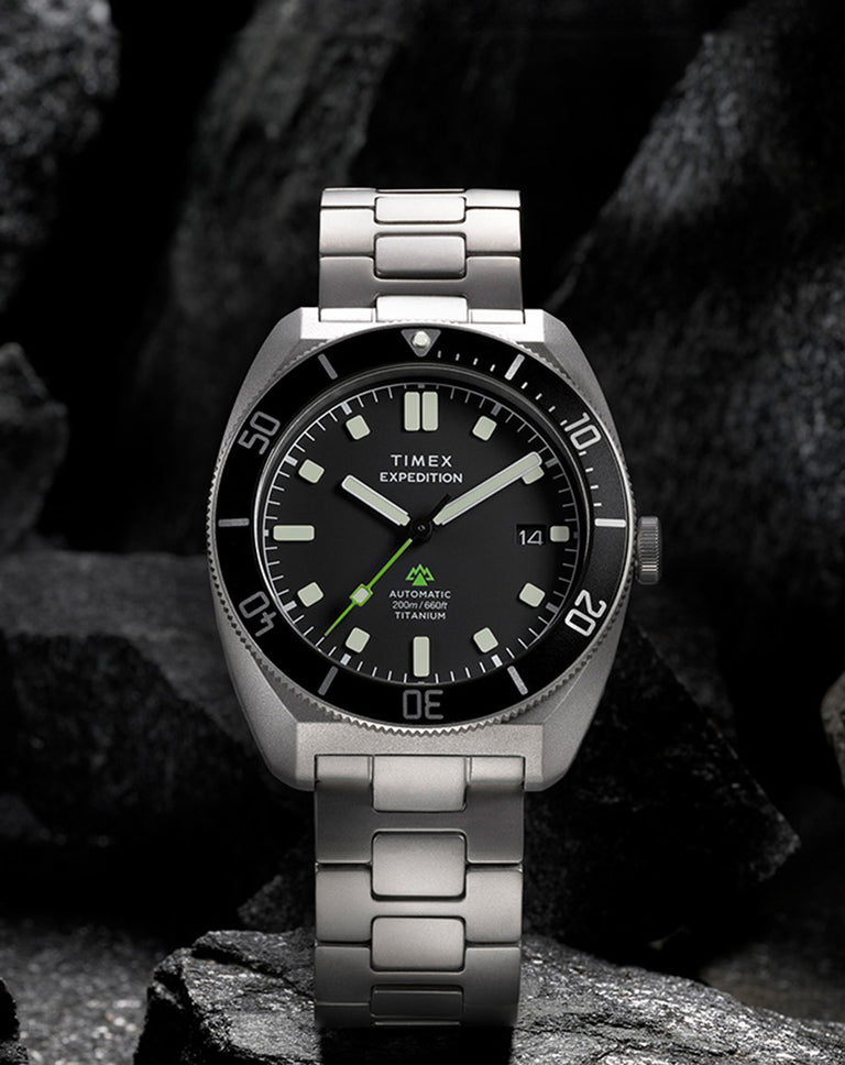 Timex Expedition Pioneer Titanium Automatic watch with a black dial and titanium bracelet displayed against a dark rocky background.