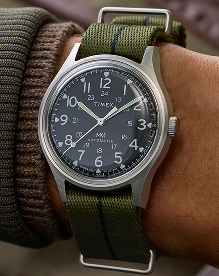 Timex MK1 Automatic watch with black dial, full Arabic numerals, and green fabric strap worn on wrist.