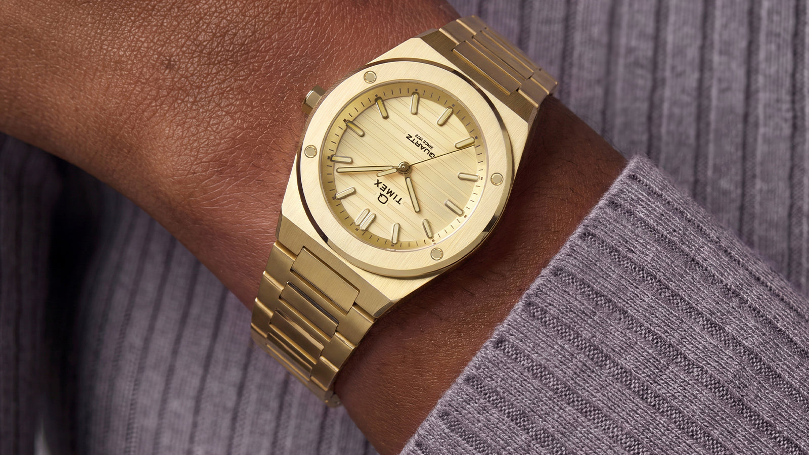 Q Timex Marbella watch with a gold-tone stainless steel case and integrated bracelet, featuring a champagne sunray dial with gold-tone hands and markers, worn on the wrist.