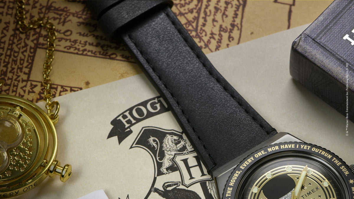 TW2Y76000 Detail of the black strap and part of the watch beside a gold Time-Turner pendant and Hogwarts-themed papers.
