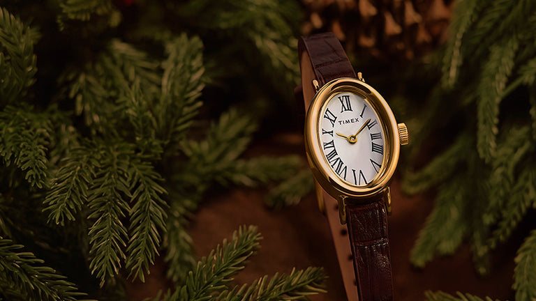 Holiday Favorites - Timex watch with an oval gold-tone case, white Roman numeral dial and brown leather strap among evergreen branches.
