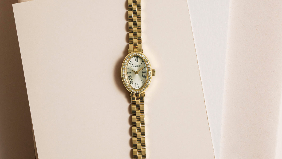 TW2Y90100 Gold-tone Timex Cavatina® Crystals watch with an oval Roman numeral dial and crystal-lined bezel, laid flat on cream paper.