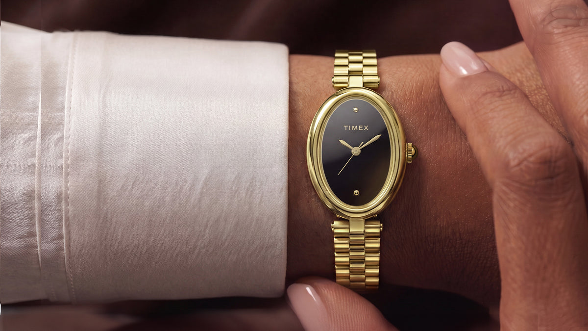 TW2Y86200 Person wearing the Timex Cavatina® Luxe gold-tone watch with a black dial while seated outdoors near the water, styled with a white outfit and sunglasses.