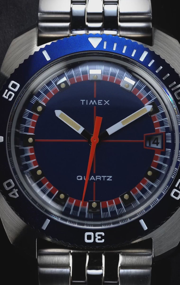 Dynamic video showcasing the Q Timex® 1971 Velocity watch. The sequence begins with high-contrast visuals of a checkered racing flag and the bold red “1971” text over a racetrack, evoking vintage motorsport themes. The camera transitions into close-up pans of the watch's navy blue dial, bright orange accents, and rotating bezel. Dramatic lighting highlights the stainless steel bracelet, angular case profile, and domed crystal. 