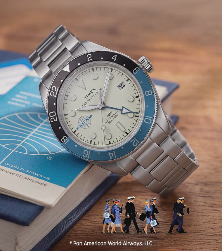 TW2Y94300 Pan Am® x Timex® Waterbury GMT watch with a cream dial, blue-and-black bezel and silver-tone bracelet resting on a Pan Am booklet, with miniature airline crew figures below and an animated airplane flying overhead.