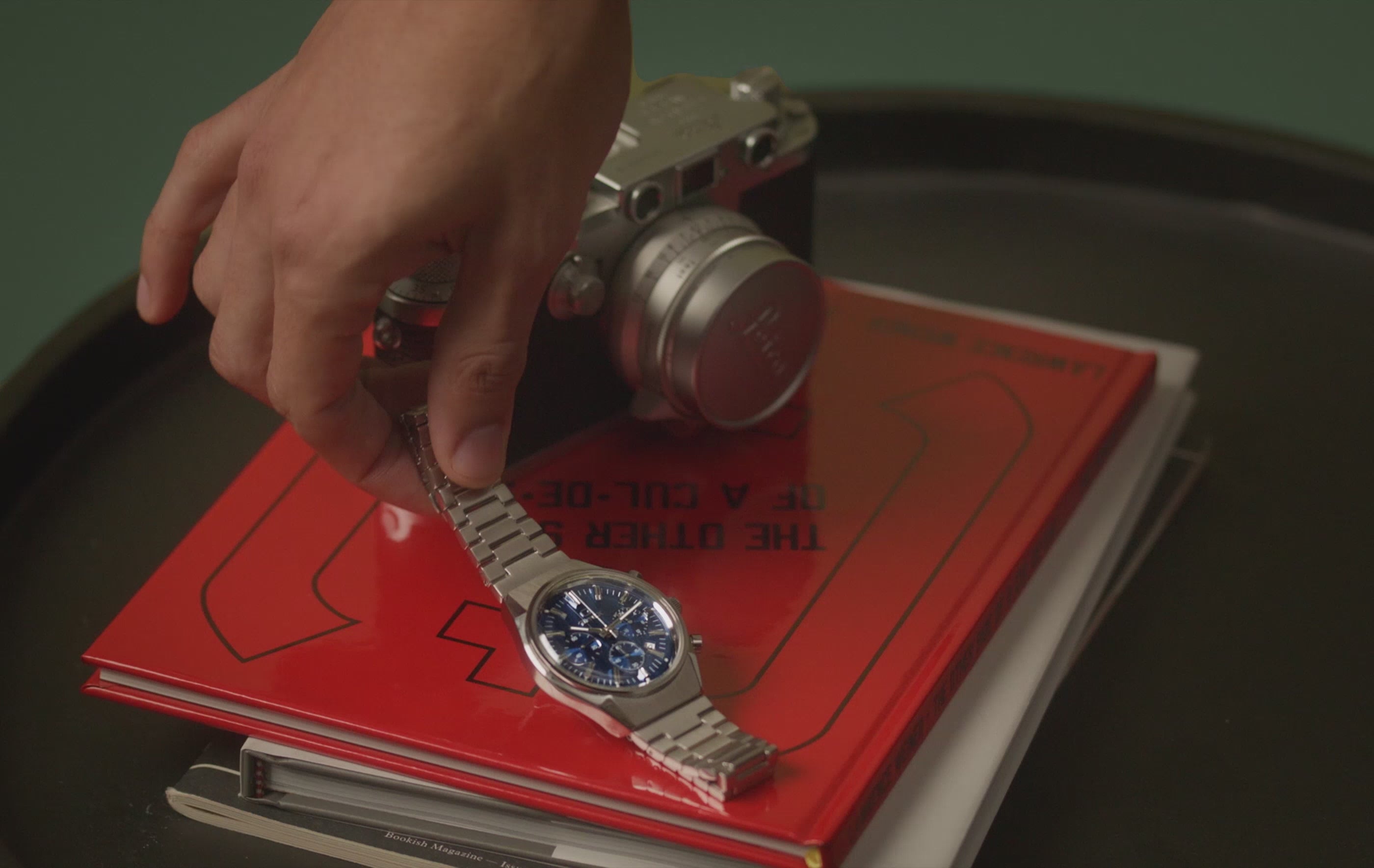 Falcon Eye Chronograph video featuring a man demonstrating putting the watch on his wrist and then laying the watch back down against a red book of Lawrence Weiner: The Other Side of A Cul-De-Sac 