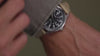 Person wearing the MK1® Automatic watch while handling binoculars, followed by close-up views of its black dial, silver-tone case, and tan fabric strap.