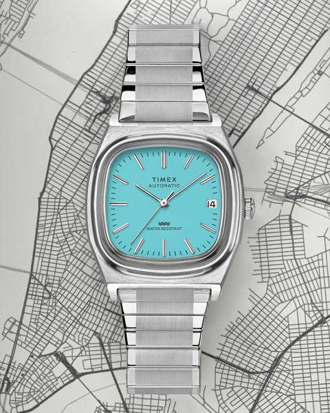 A Timex automatic watch featuring a striking turquoise dial with a date display, encased in a brushed stainless steel case and bracelet. The background showcases a detailed black-and-white map design, adding a sophisticated urban aesthetic.